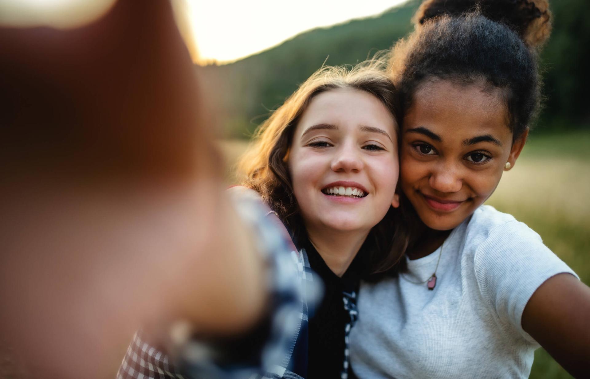 Two smiling young women taking a selfie in a scenic outdoor setting. The mood is joyful and carefree, with greenery blurred in the background.
