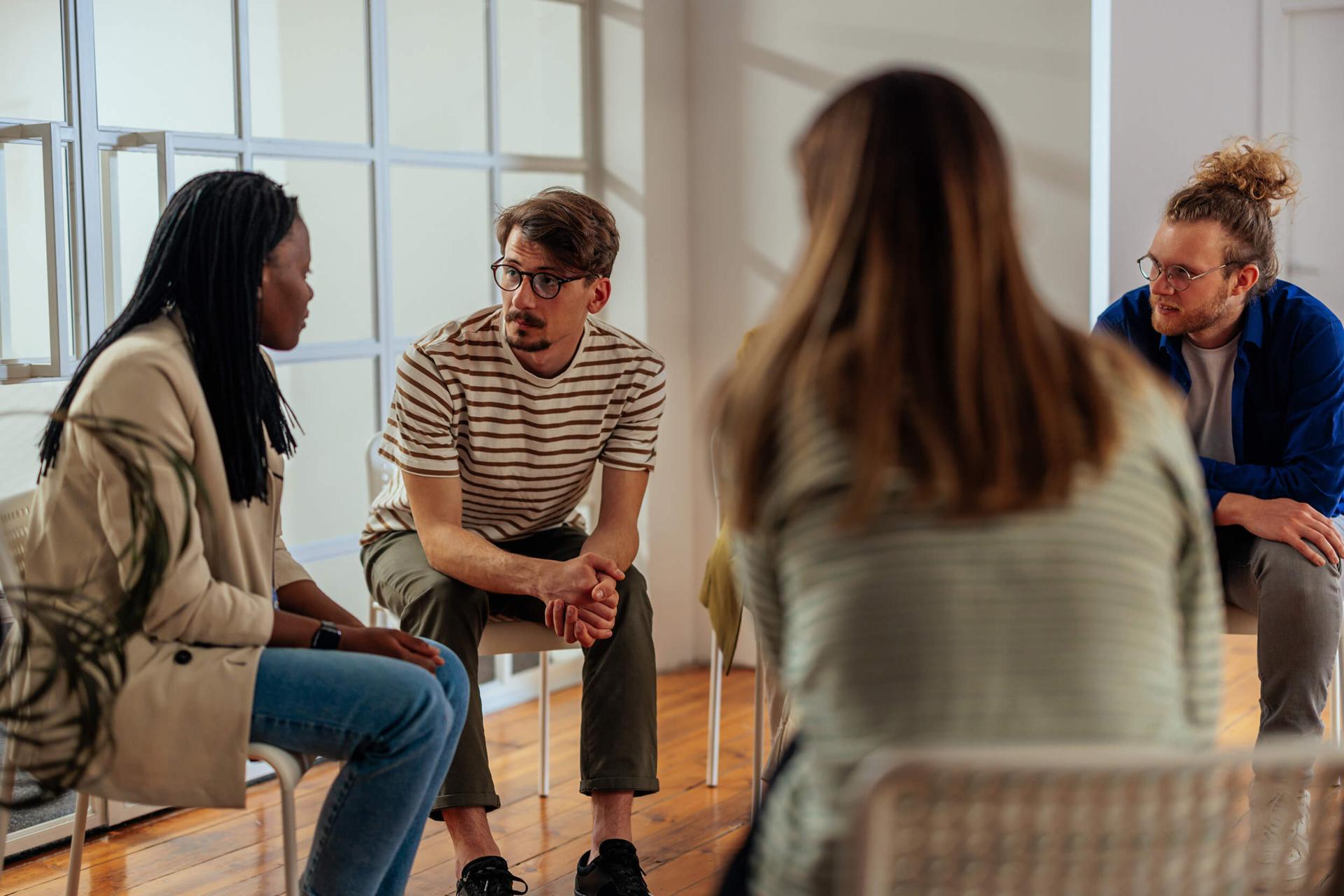 A diverse group of four people sits in a circle in a bright room, engaged in a serious discussion. Their body language conveys focus and attentiveness.