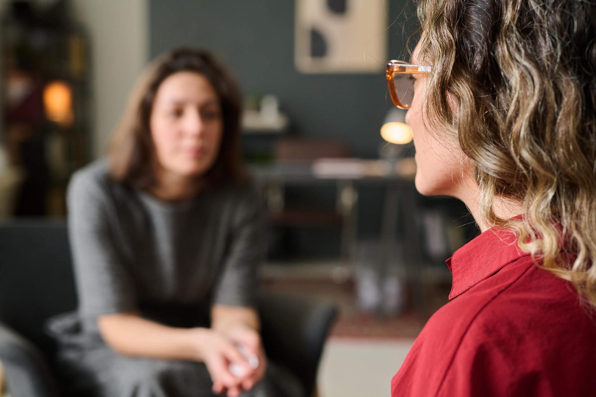 A woman with curly hair and glasses listens attentively to another woman in a blurred background, suggesting a supportive and conversational setting.