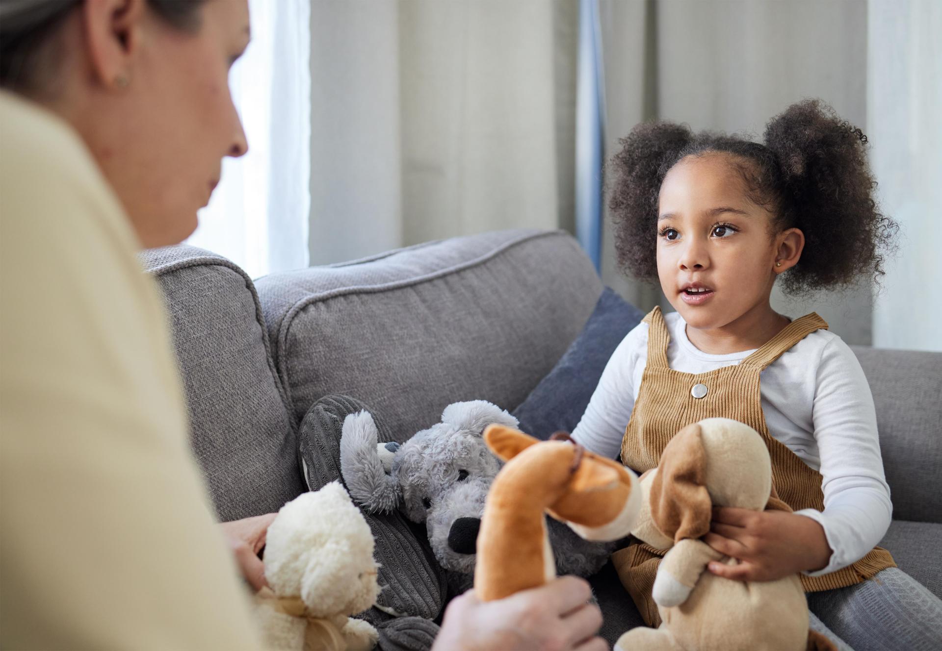 A young girl with curly hair, wearing a brown dress, sits on a couch with stuffed animals, attentively interacting with an adult. The scene is cozy and engaging.