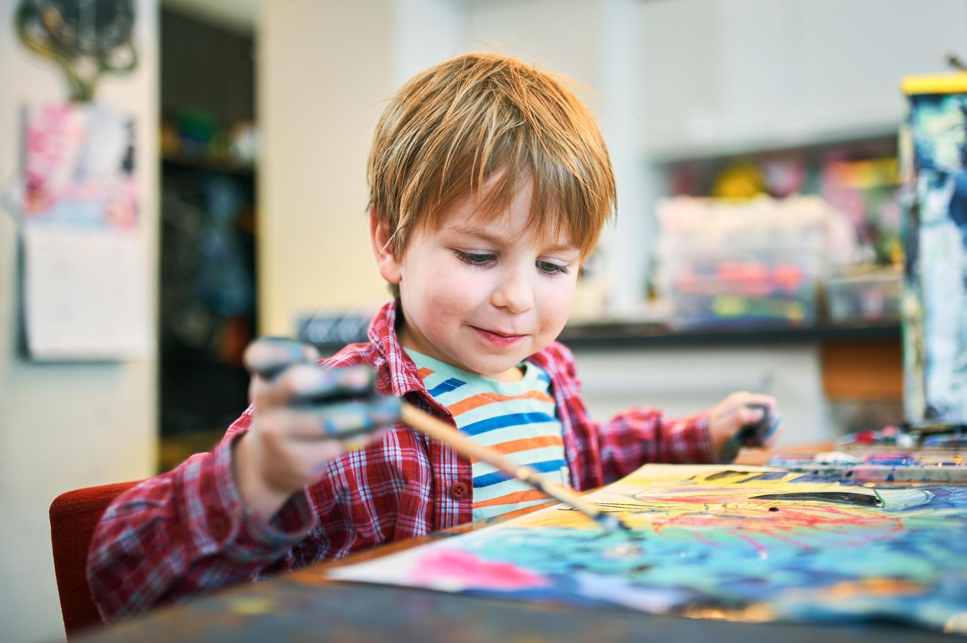 A young boy with light brown hair paints with vibrant colors on paper. He wears a red plaid shirt and smiles, showing joy and creativity in a home setting.