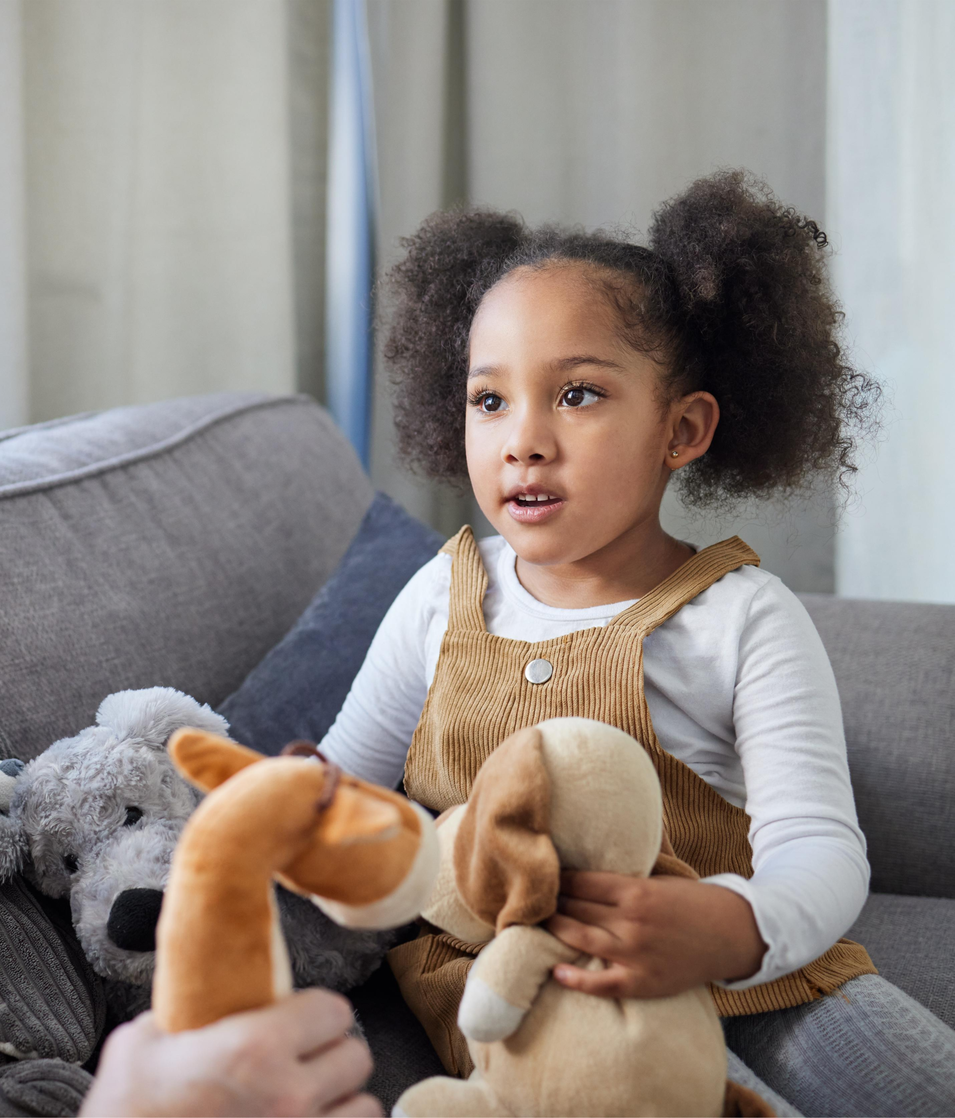 A young girl with curly hair, wearing a brown dress, sits on a couch with stuffed animals, attentively interacting with an adult. The scene is cozy and engaging.