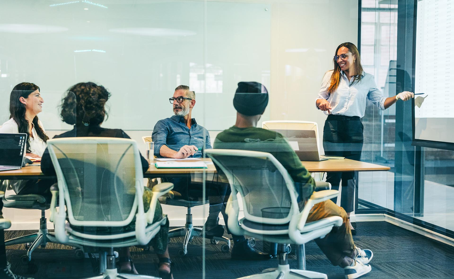 A diverse team in a meeting room engaged in a presentation. A smiling woman points to a screen, creating a collaborative and positive atmosphere.