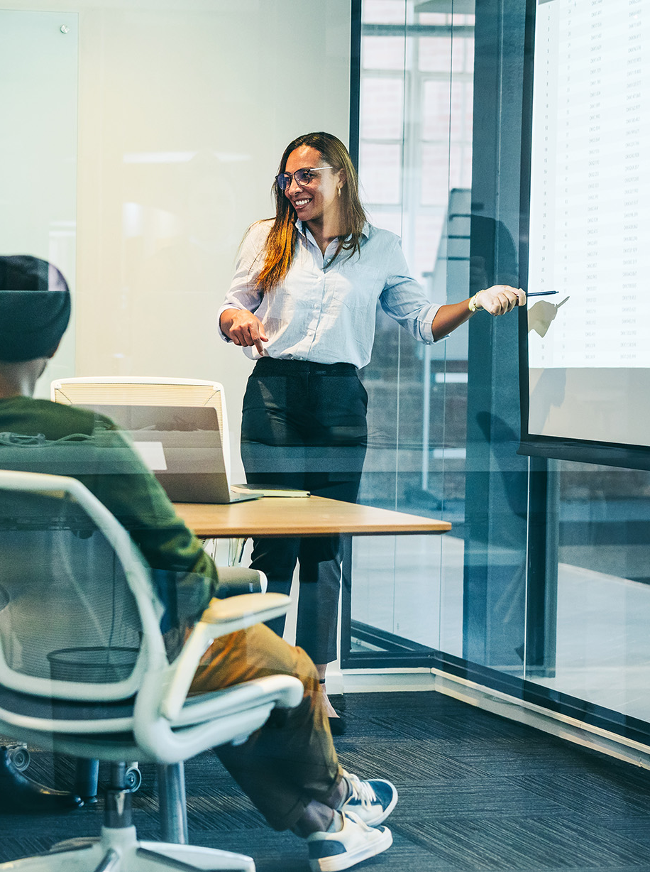 A diverse team in a meeting room engaged in a presentation. A smiling woman points to a screen, creating a collaborative and positive atmosphere.