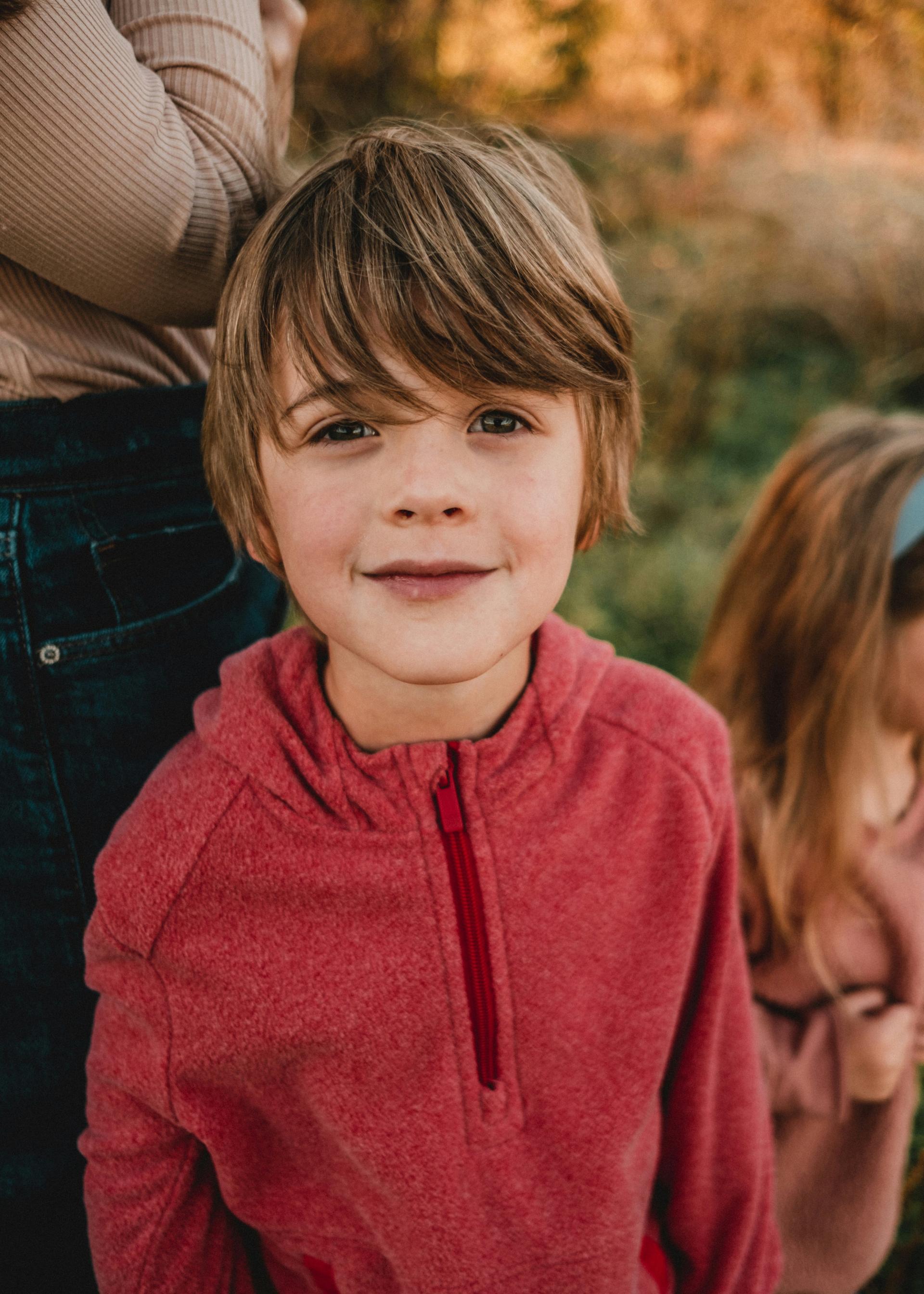 Young boy in a red sweater smiles warmly at the camera, standing outdoors next to a blurred figure. Autumn tones create a cozy, joyful atmosphere.