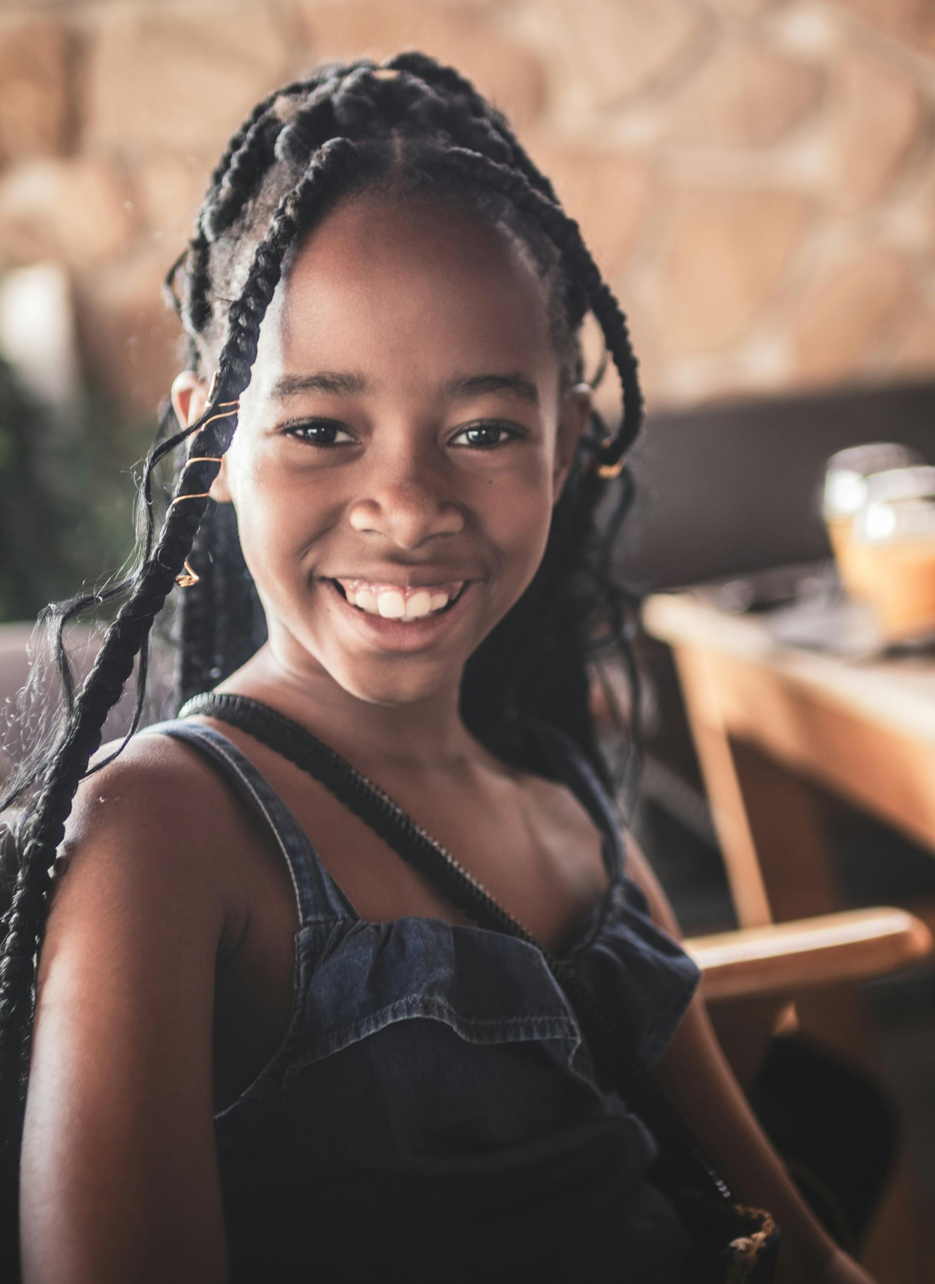 Smiling young girl with braided hair sits indoors, wearing a blue dress. The background is softly blurred, creating a warm, cheerful atmosphere.