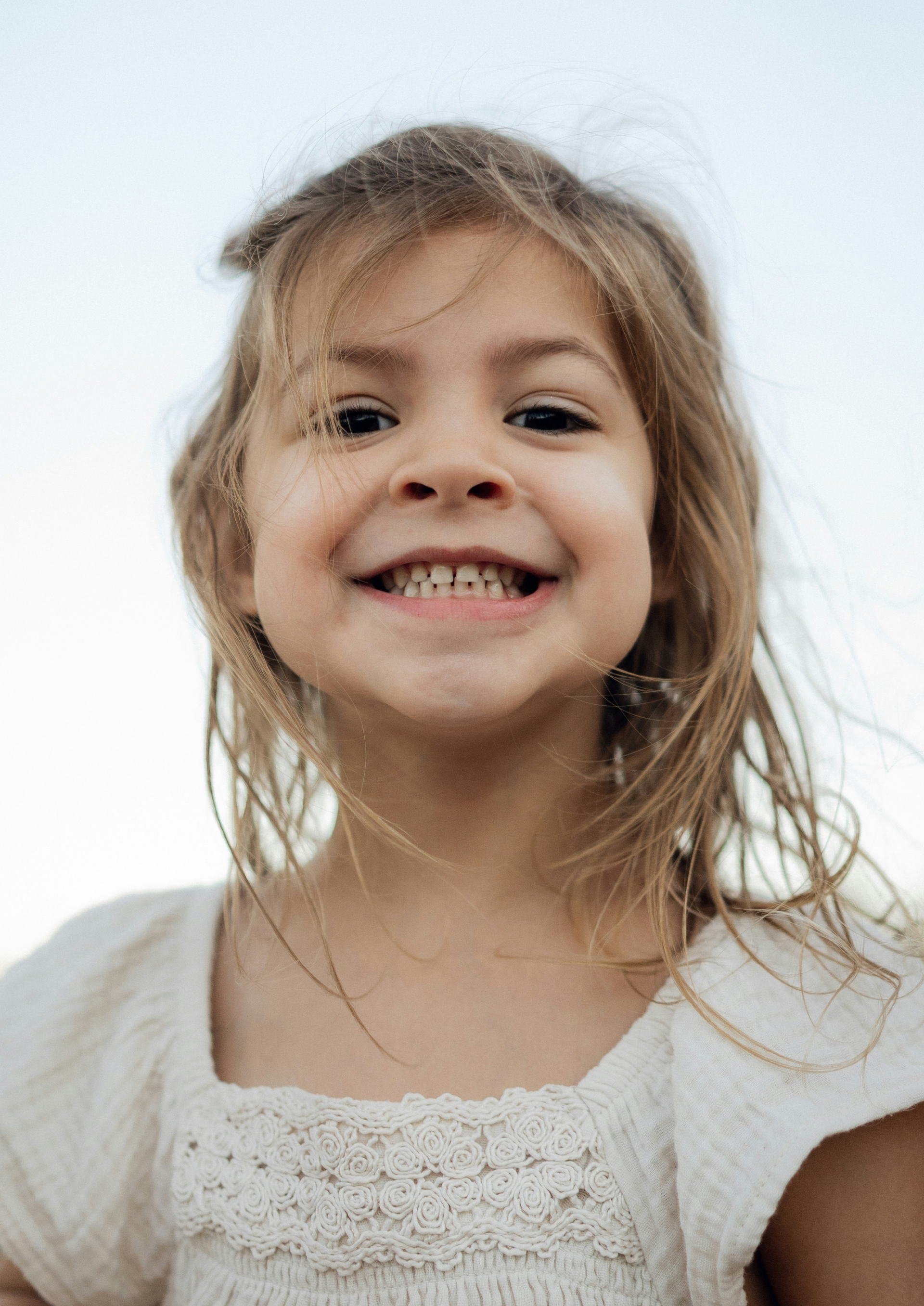A young child with messy hair smiles widely at the camera, wearing a white dress against a clear blue sky. The scene conveys joy and innocence.