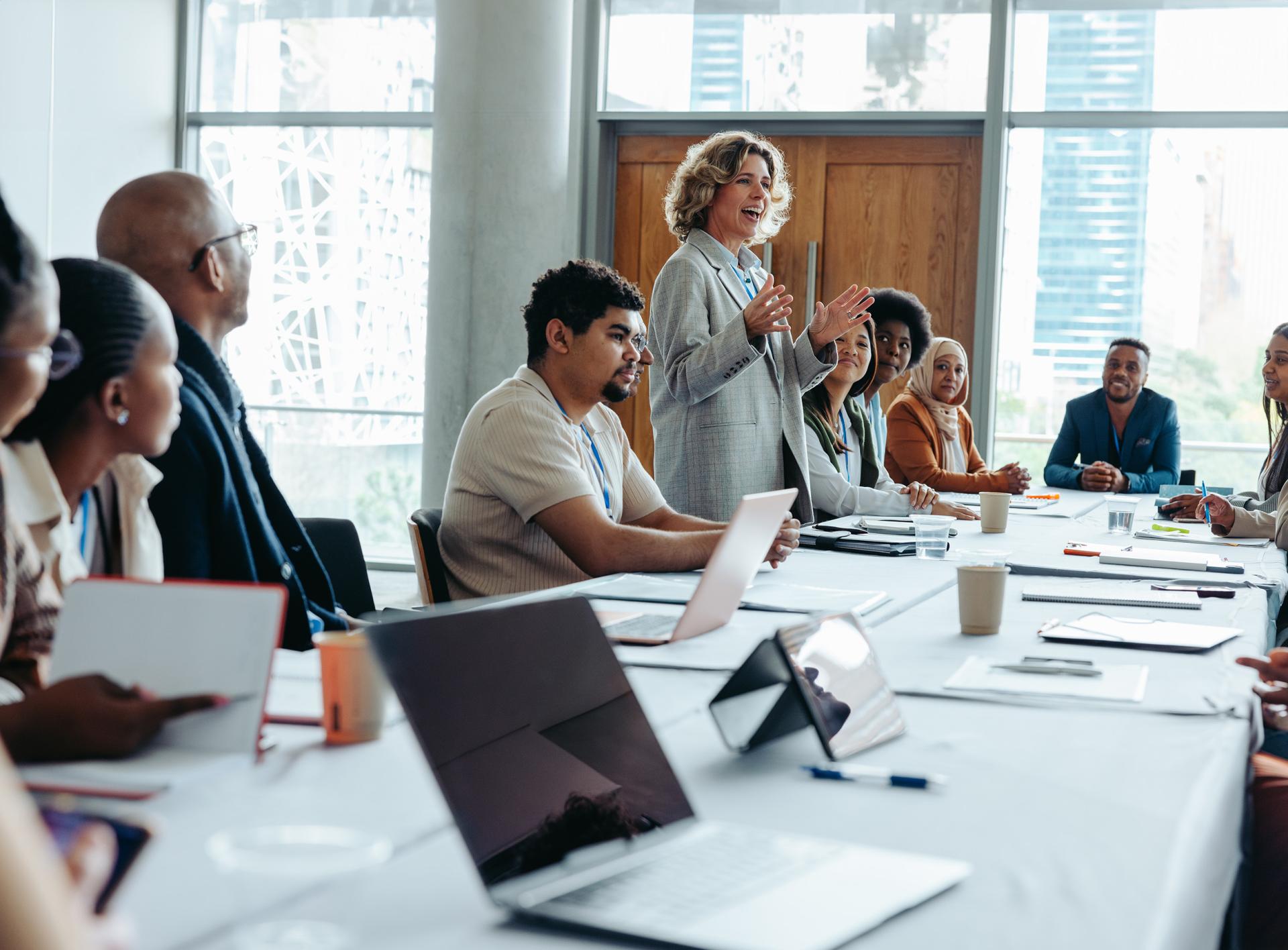 A diverse group of professionals sit around a conference table. A woman stands, speaking confidently, suggesting an engaging business meeting. Laptops and notepads are visible.