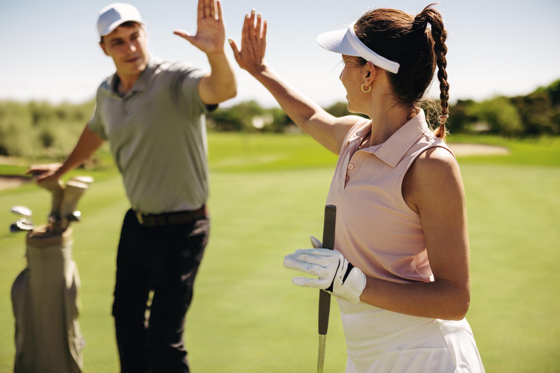 A man and woman share a high-five on a sunny golf course, holding clubs and wearing visors. Their expressions show joy and camaraderie.