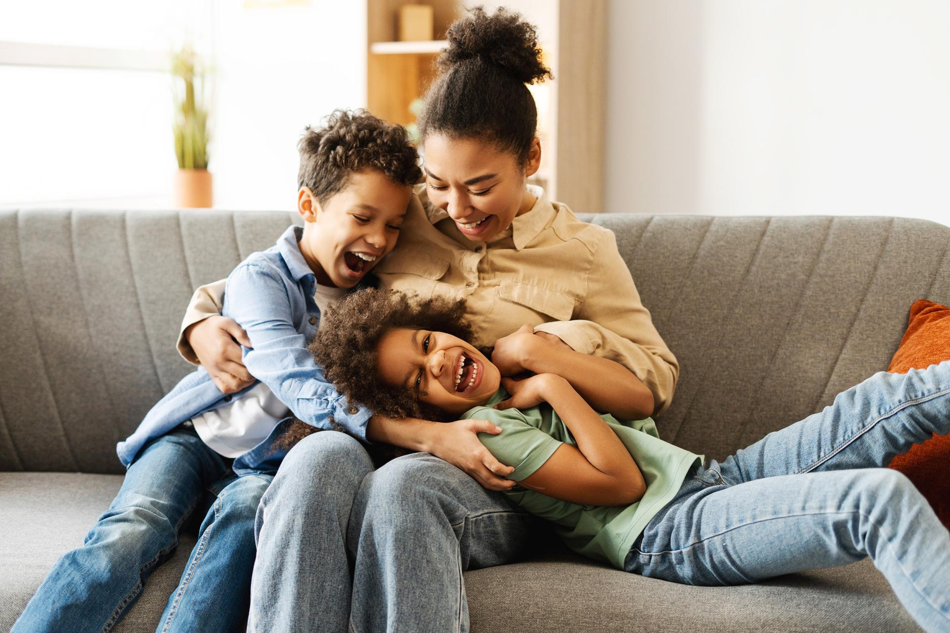 A joyful mother sits on a gray couch, hugging and laughing with her two young children. The scene is warm and cheerful, with a cozy home ambiance.