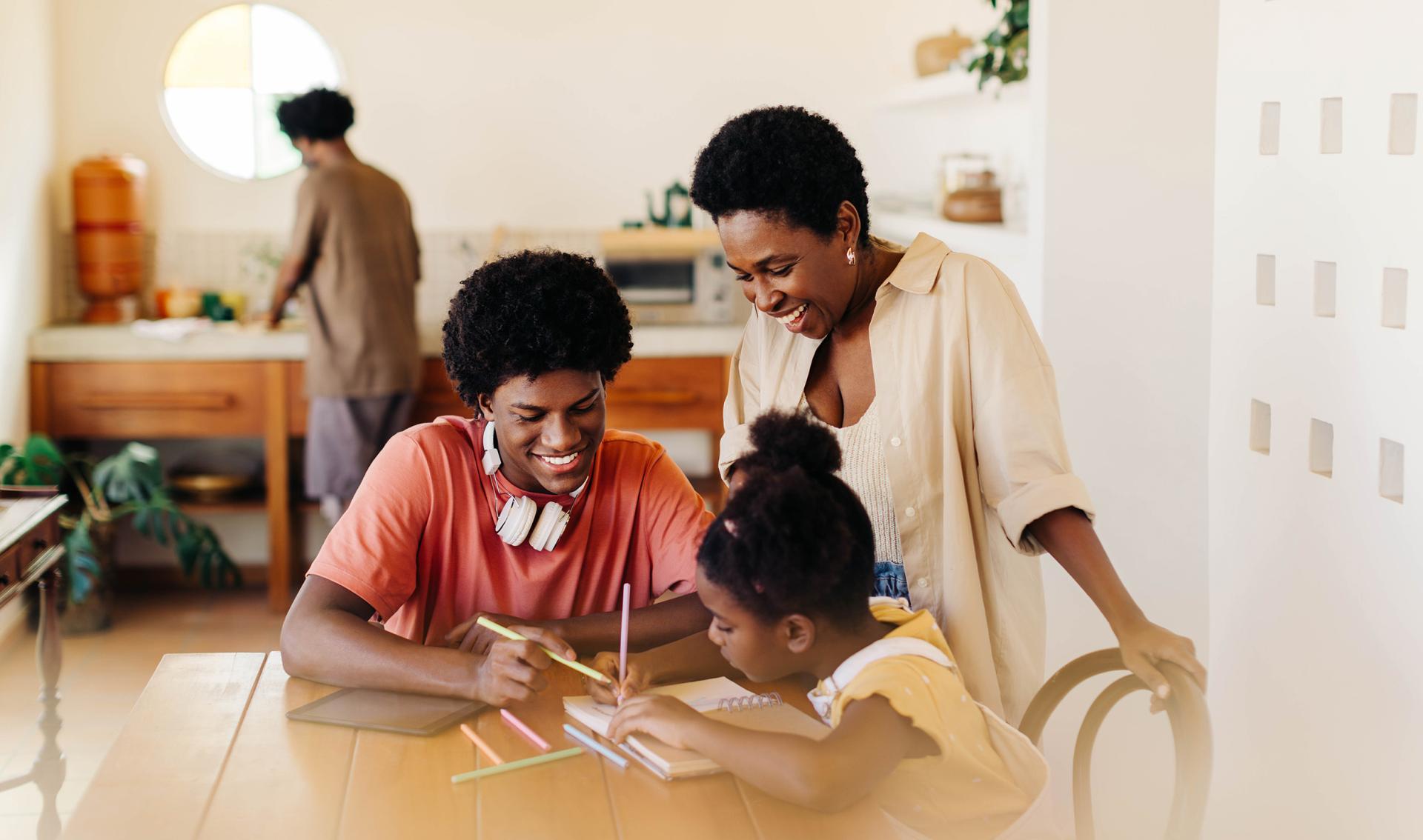 A family gathers around a wooden table in a bright kitchen. A smiling woman watches a teenage boy and young girl draw with colored pencils. A man is in the background.