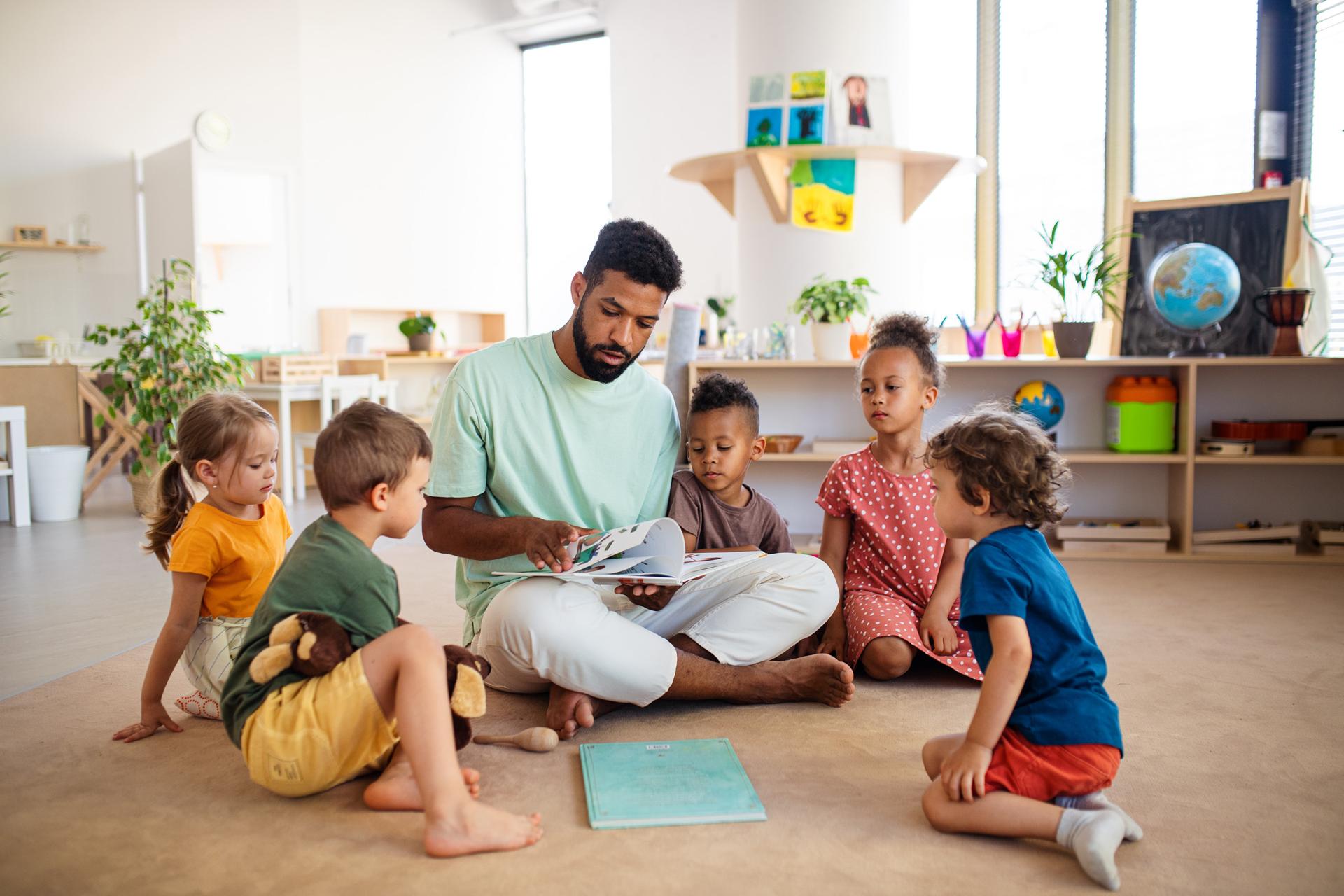 A teacher sits cross-legged on the floor, reading a book to five young children. The kids are seated around him, listening intently in a classroom setting.