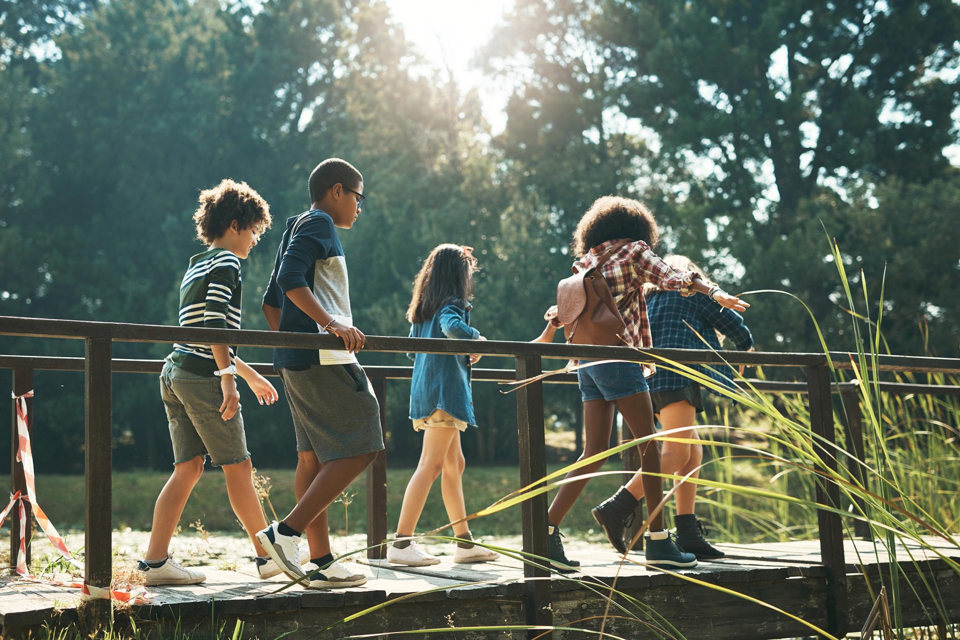 A group of five children walks across a wooden footbridge in a sunlit park. They appear joyful and adventurous, surrounded by tall grass and trees.