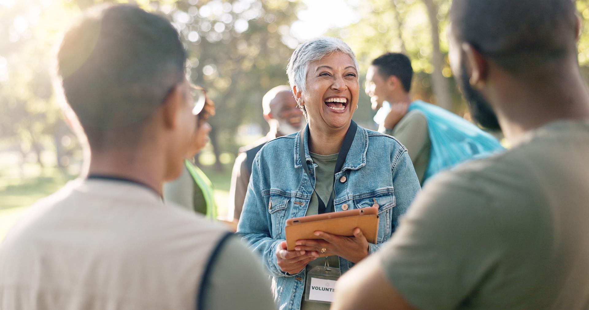 A woman in a denim jacket joyfully smiles while holding a tablet, surrounded by a group of diverse people outdoors. The setting is warm and sunny, conveying unity and positivity.
