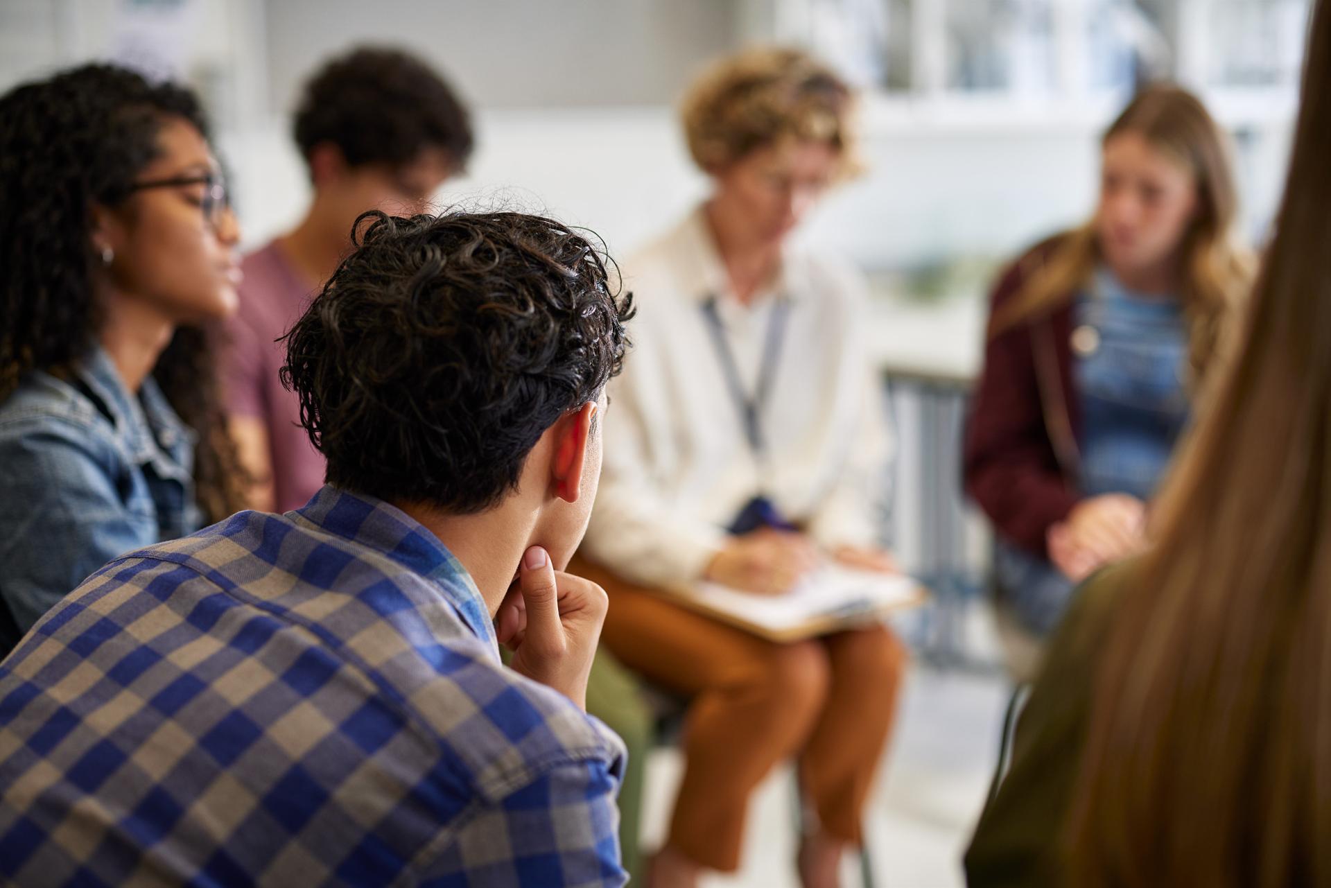 A diverse group of people sit in a circle, engaged in discussion. The focus is on a young man listening intently, creating a thoughtful atmosphere.