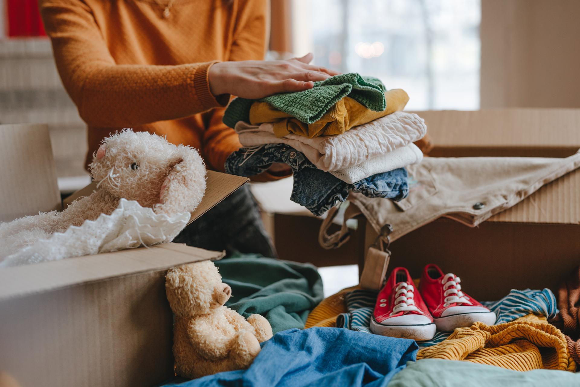 A person packs clothes into a cardboard box alongside a teddy bear and sneakers. The scene conveys a sense of organization and preparation.