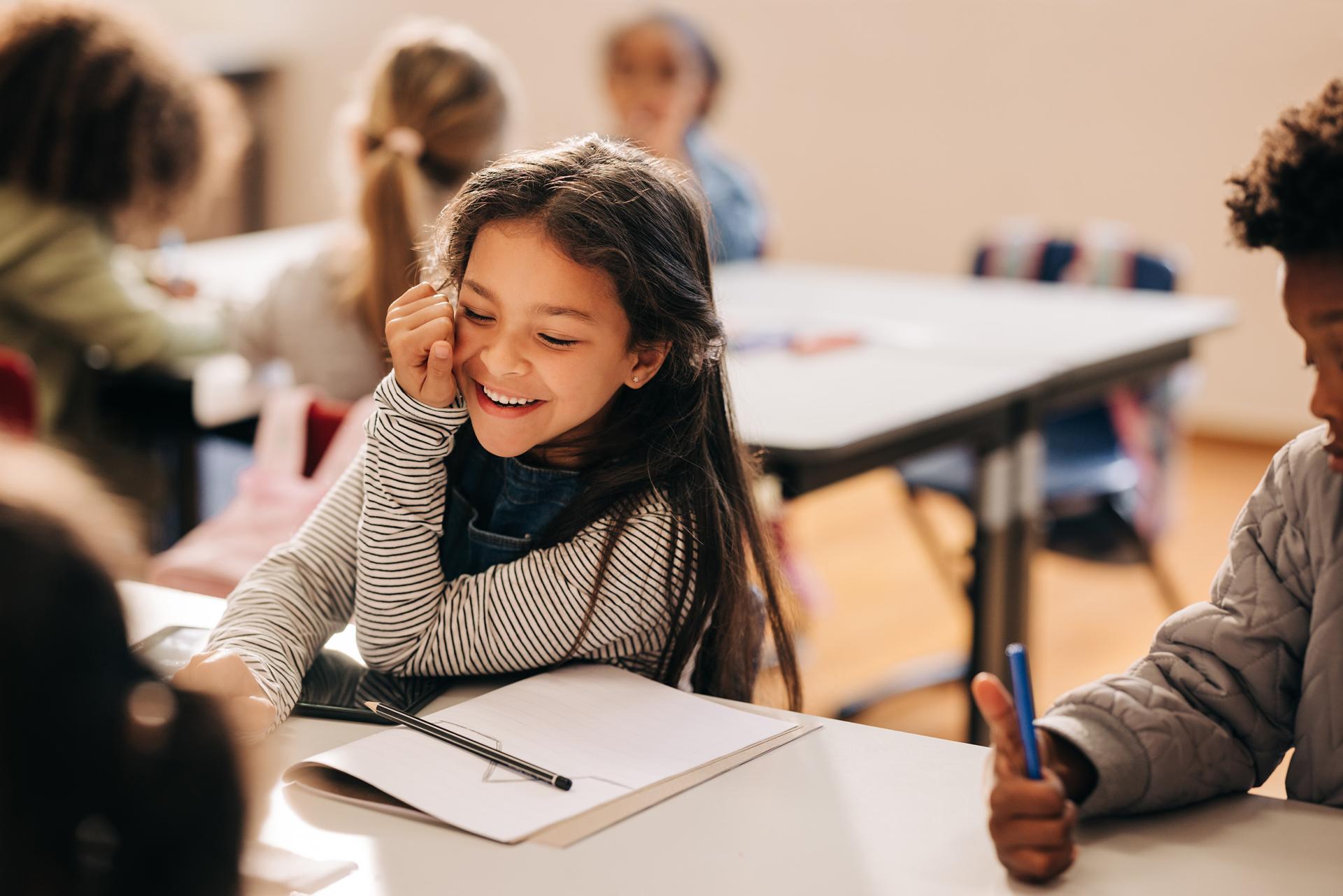 A smiling young girl seated at a classroom table with a notebook and pen in front of her.