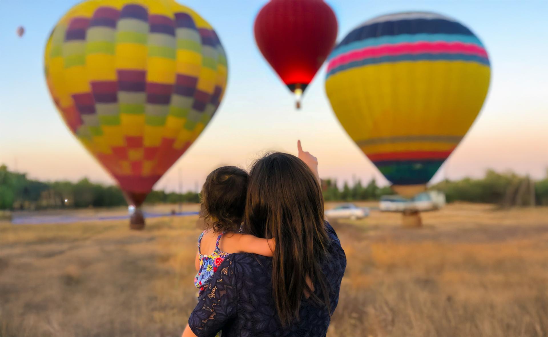 A woman holds a child, pointing towards three colorful hot air balloons in the sky at sunset. The scene conveys wonder and excitement.