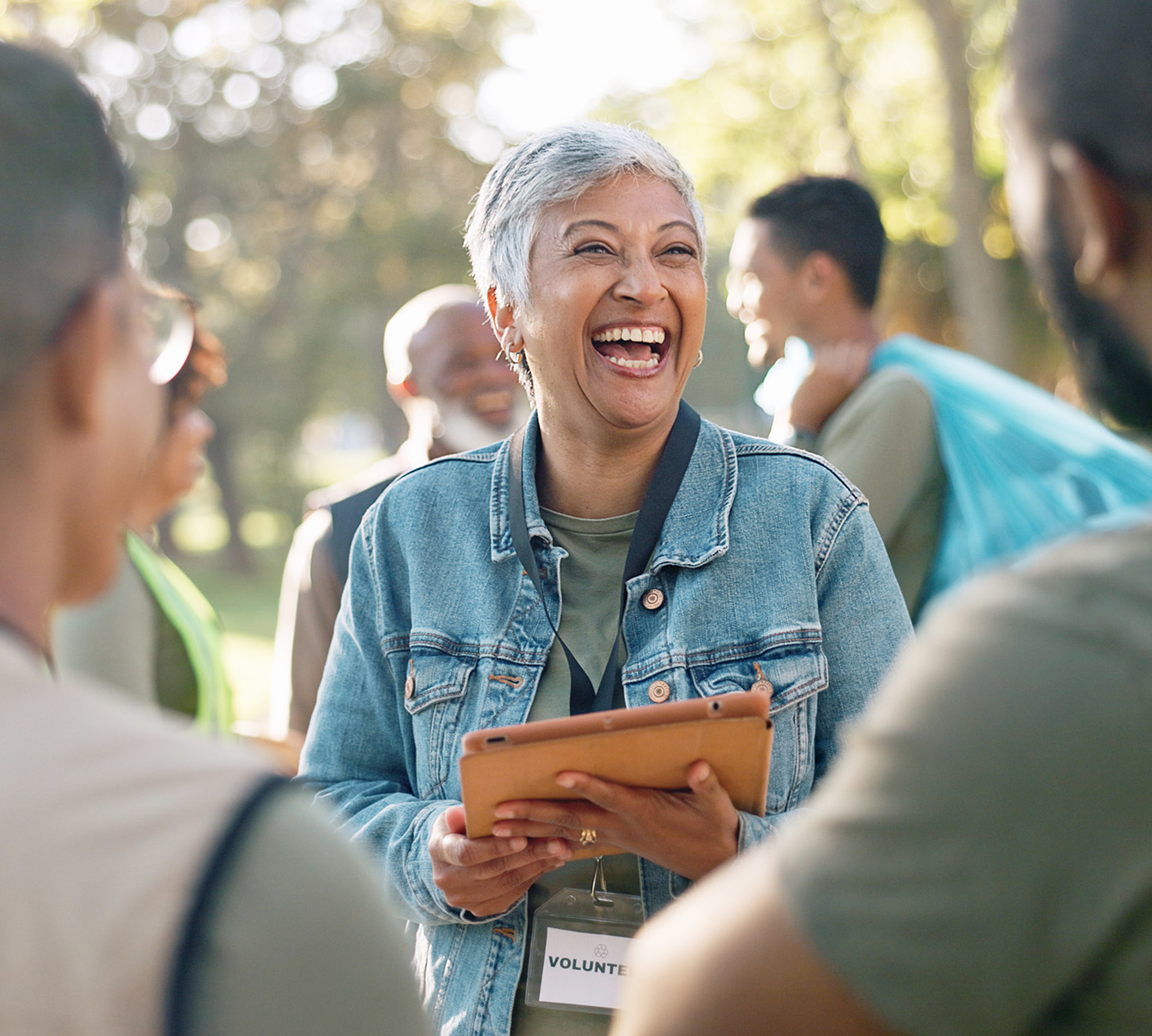 A woman in a denim jacket joyfully smiles while holding a tablet, surrounded by a group of diverse people outdoors. The setting is warm and sunny, conveying unity and positivity.