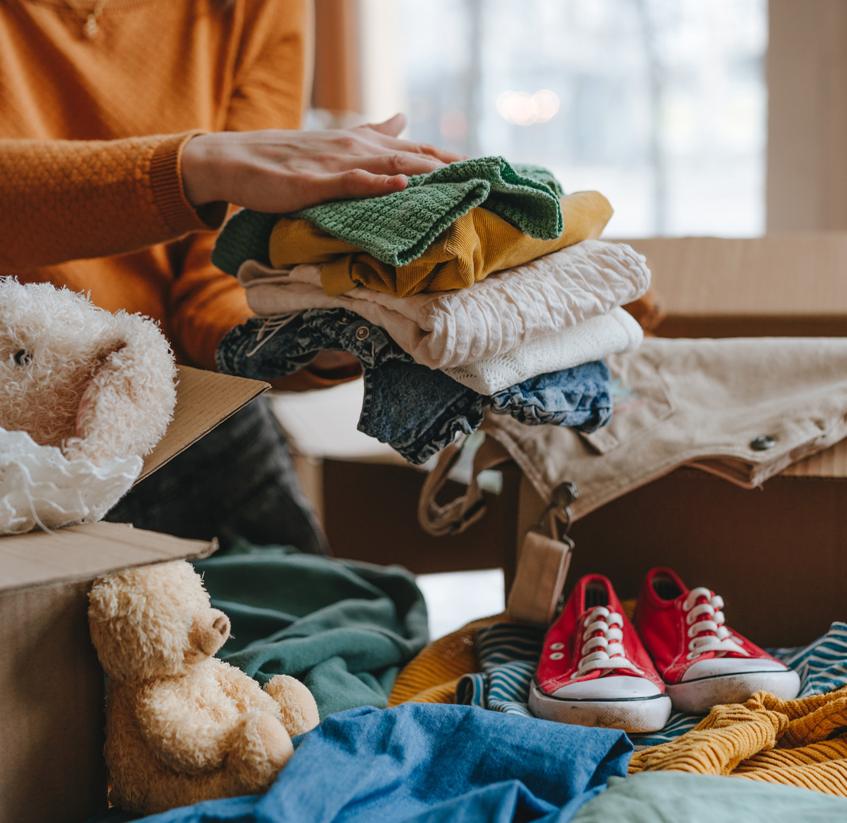 A person packs clothes into a cardboard box alongside a teddy bear and sneakers. The scene conveys a sense of organization and preparation.
