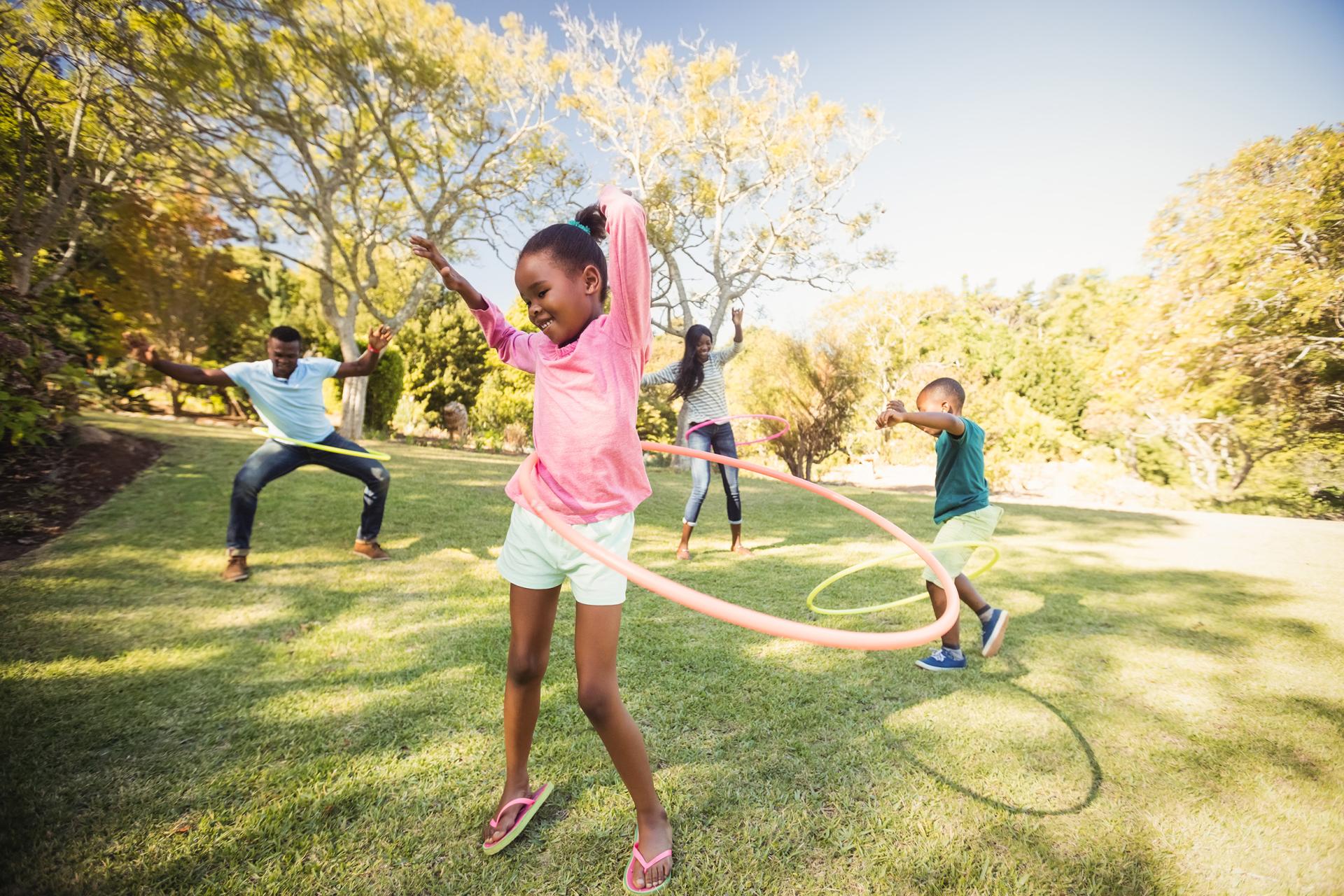 A joyful family playing with hula hoops on a sunny lawn. A girl in a pink shirt spins, while others join in the fun. Bright, cheerful atmosphere.
