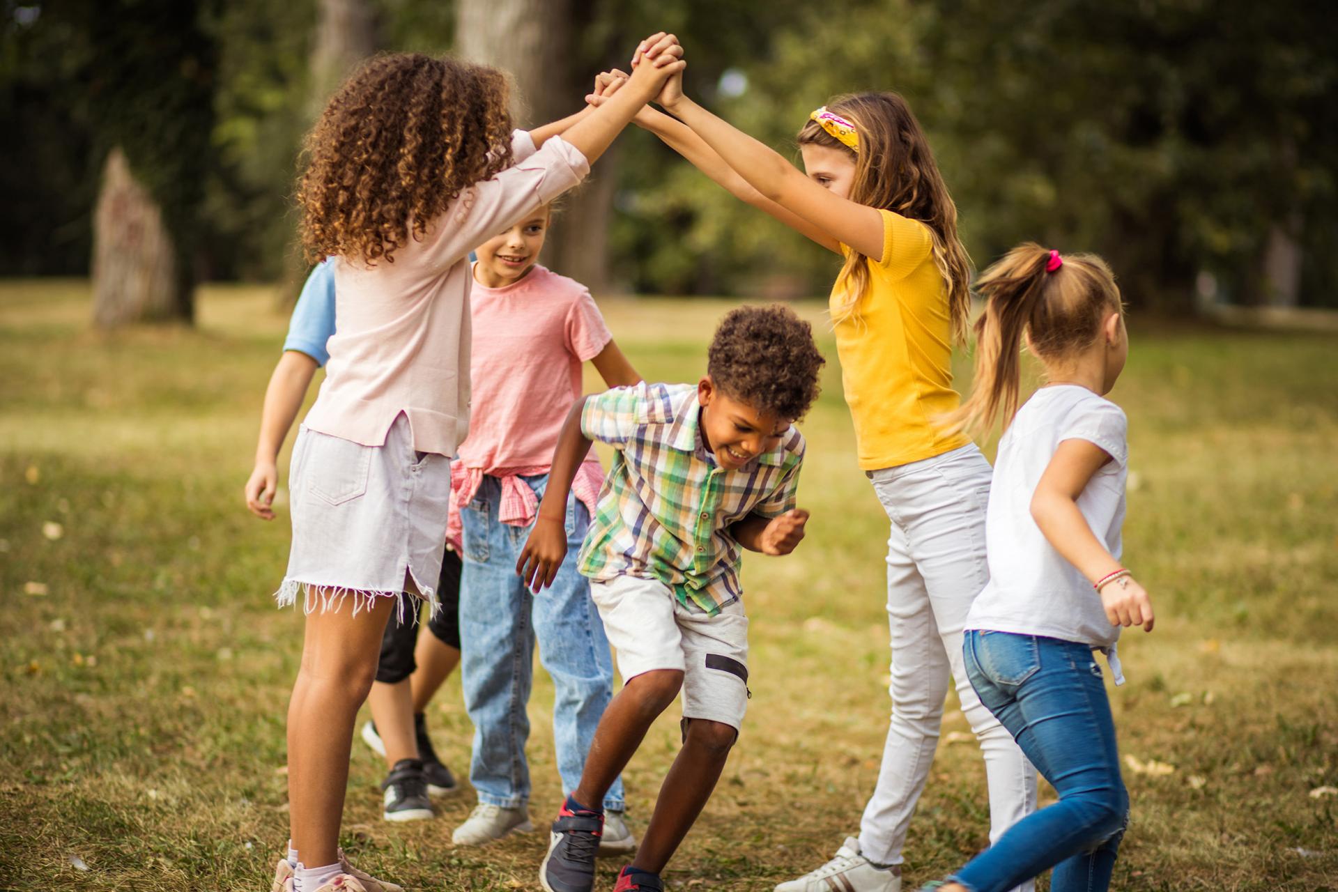 Children play a joyful game outside, forming a tunnel with raised arms. A boy in a plaid shirt ducks through, surrounded by smiling friends.