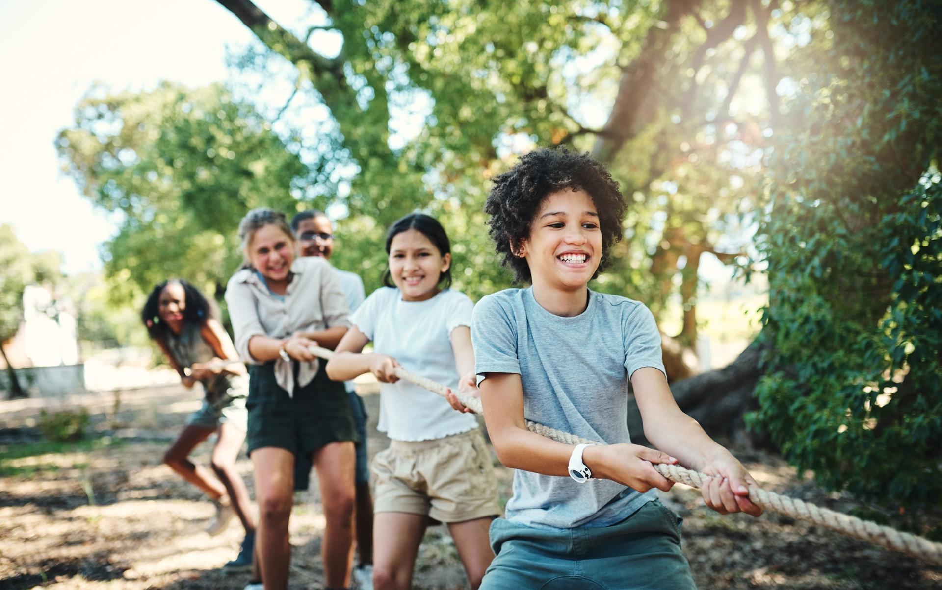 A group of smiling children play tug-of-war in a sunlit park, pulling a rope with effort and joy. Lush green trees create a lively backdrop.