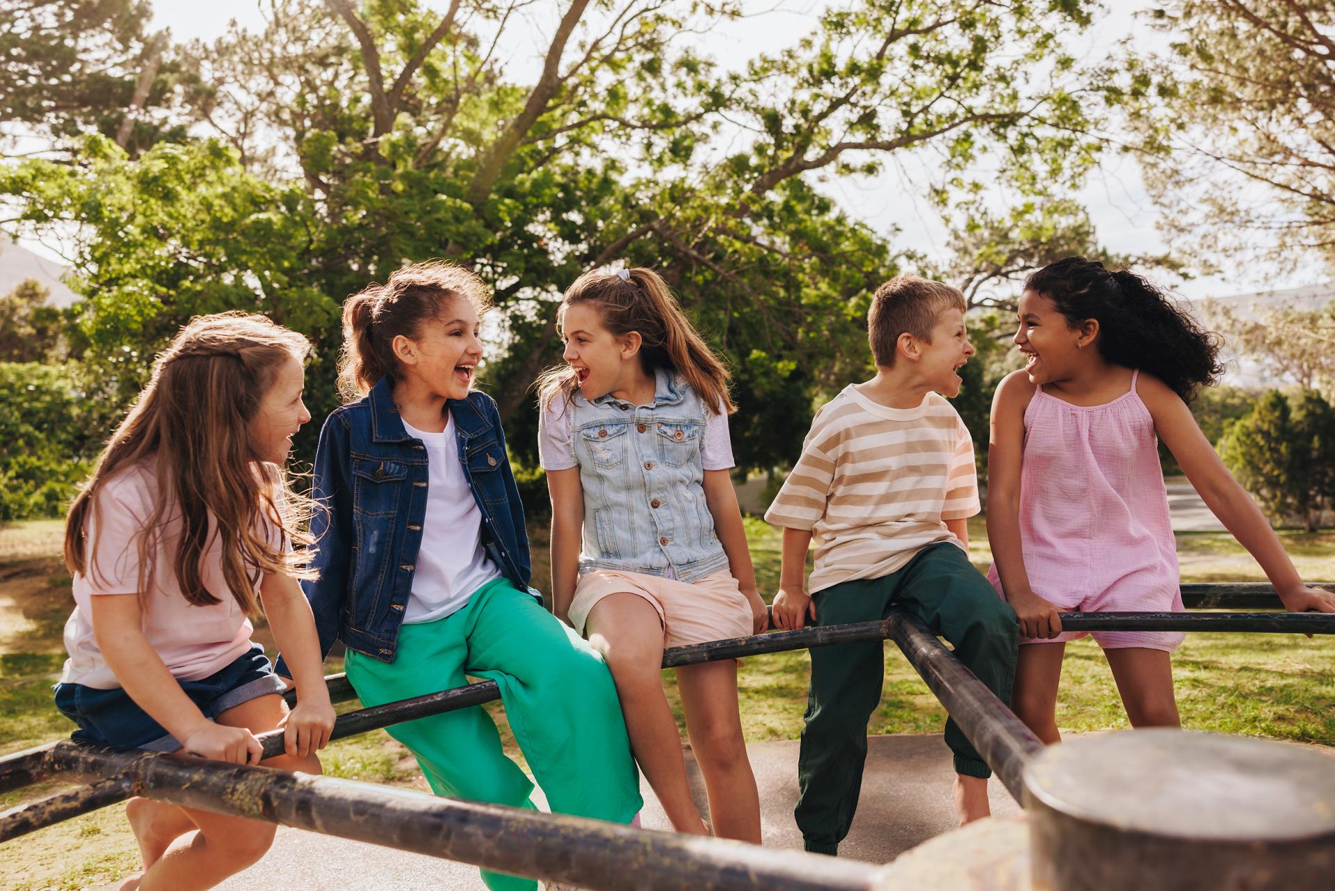 Five children smiling and sitting on a playground structure, surrounded by lush trees and sunlight. The mood is joyful and playful.