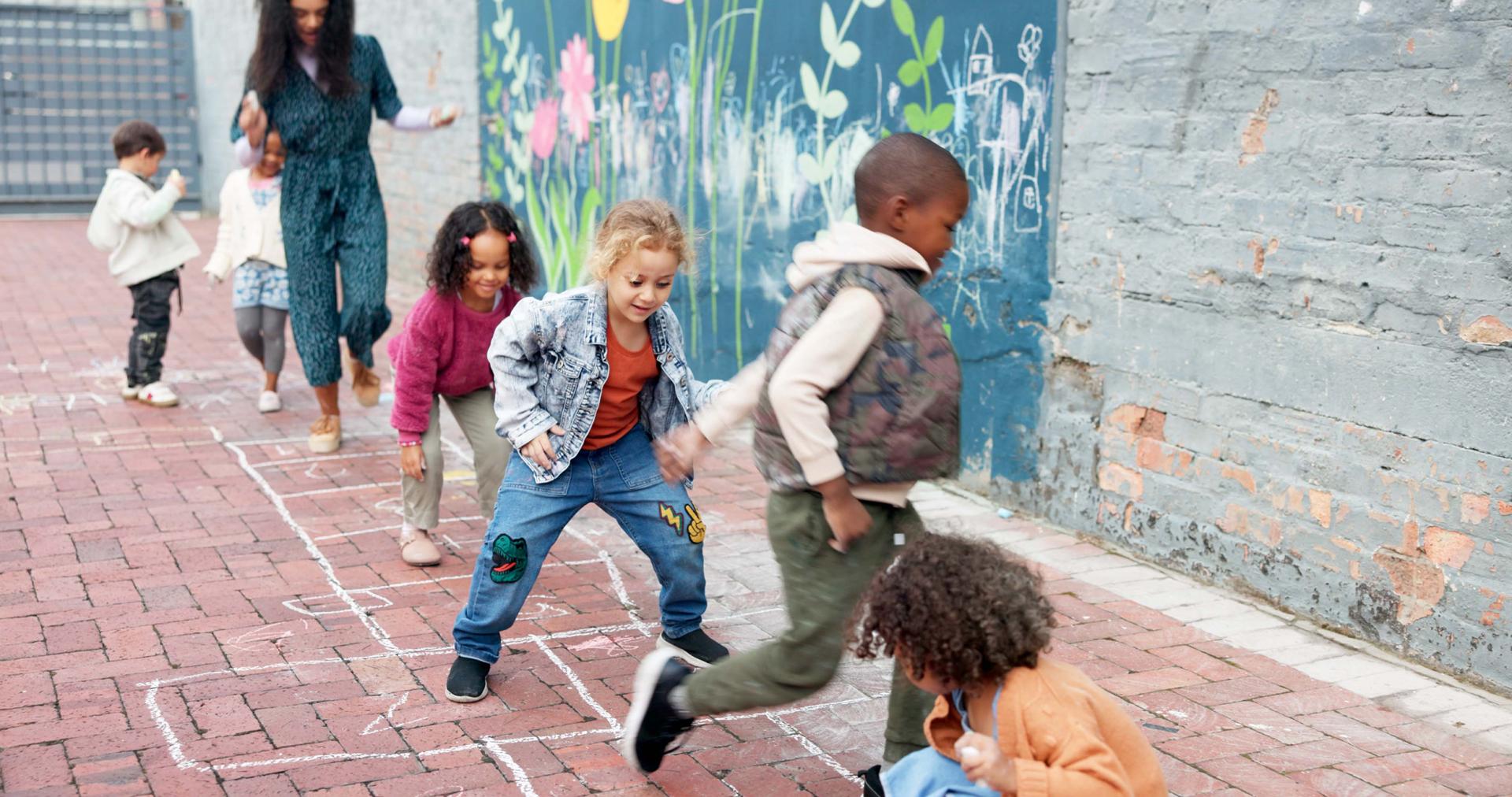 Children play hopscotch on a brick path next to a vibrant, flower-painted wall. They are joyful and energetic, led by an adult in a flowing dress.