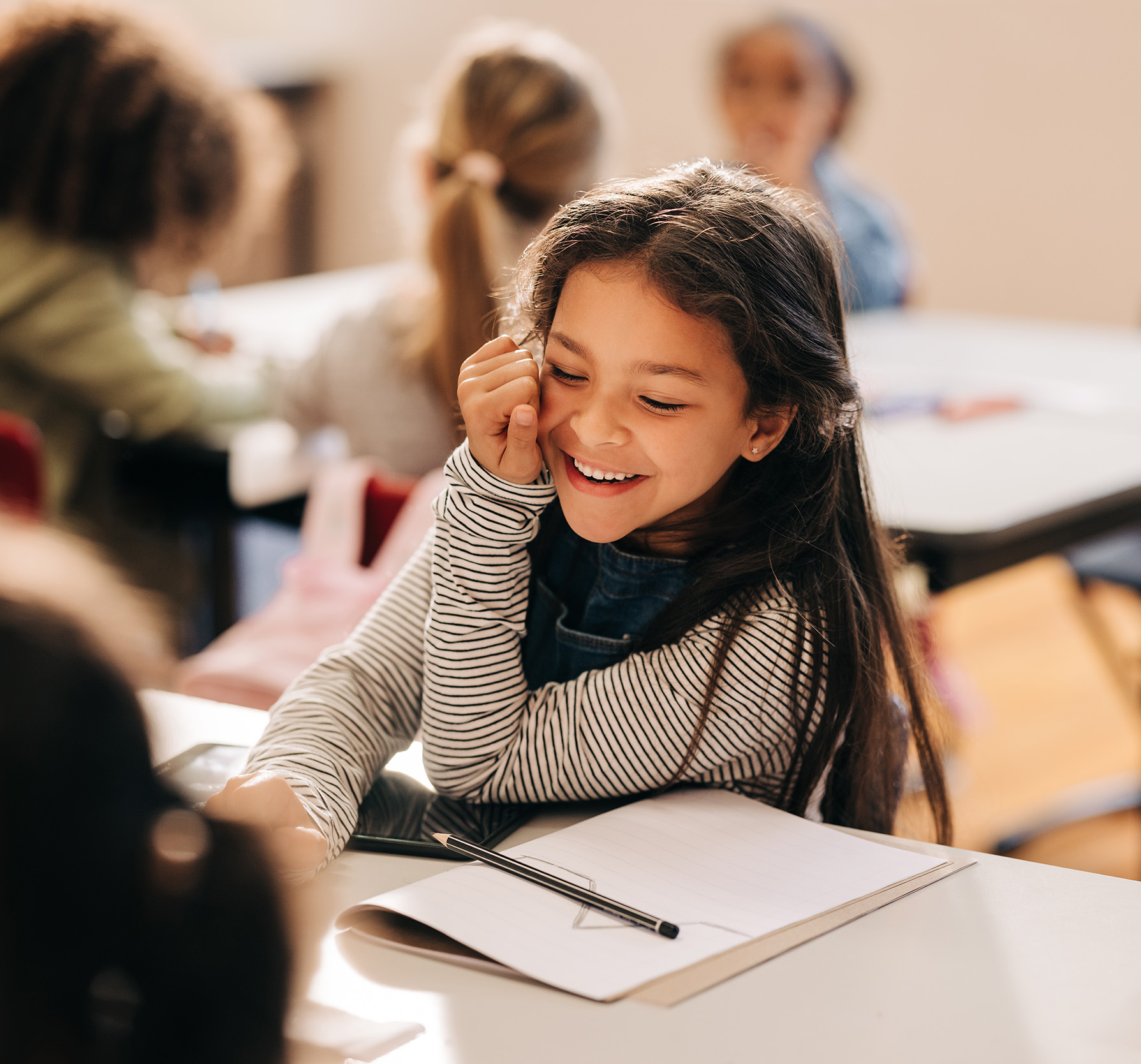 A smiling young girl seated at a classroom table with a notebook and pen in front of her.
